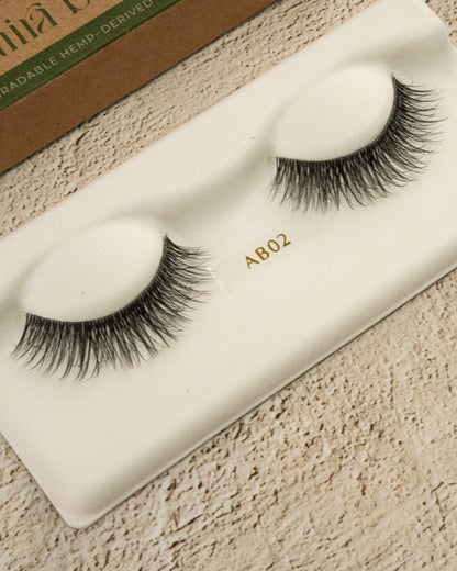 Pair of false eyelashes on a white card with 'AB02' branding, on a textured beige surface.